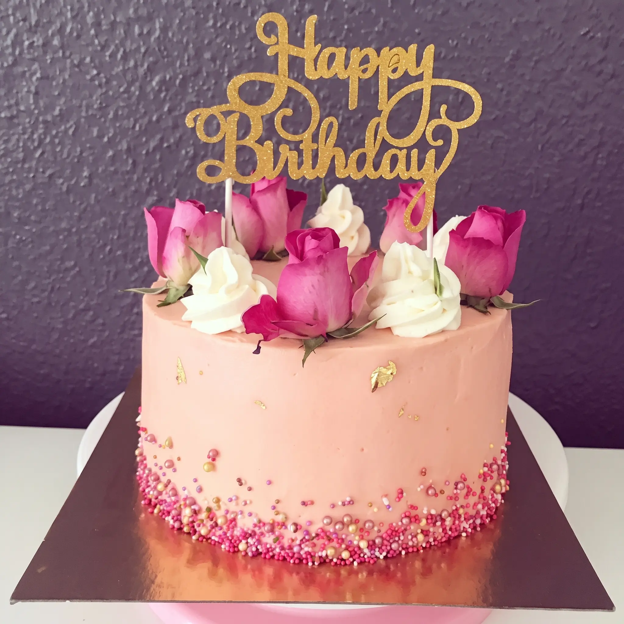 Pink birthday celebration cake decorated with roses, cream swirls, gold topper and pearl details